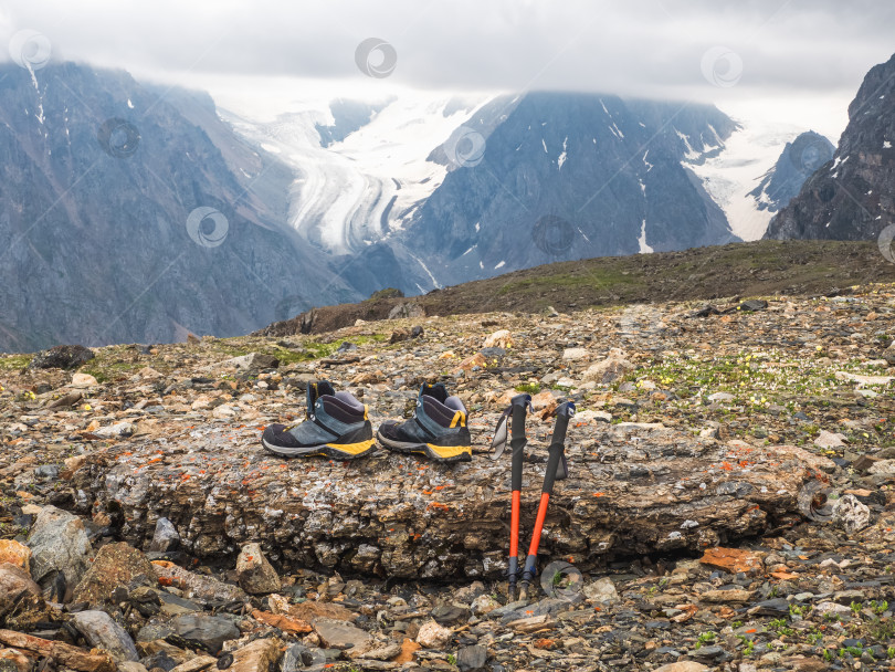 Скачать Поношенные туристические ботинки и треккинговые палки покоятся на скале на фоне ледника и высоких гор. Альпийский треккинг. фотосток Ozero