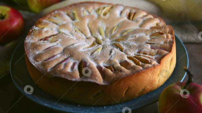 Скачать Свежеиспеченный яблочный пирог, украшенный кусочками яблок и сахарной пудрой, лежит на бирюзовой тарелке на деревянном столе в деревенском стиле в окружении спелых яблок, создавая уютную осеннюю атмосферу фотосток Ozero