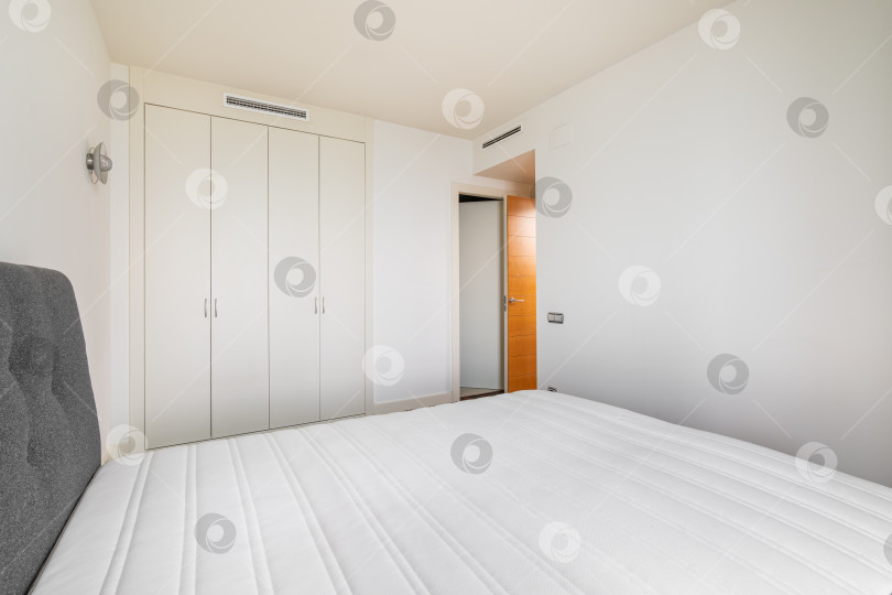 Скачать Современная светлая спальня с кроватью и встроенным шкафом-купе и открытой дверью в коридор. Минималистичная, но уютная концепция дизайна. Дизайн отеля или съемной квартиры фотосток Ozero