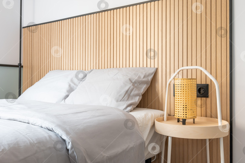 Скачать Крупным планом спальня с двуспальной кроватью на фоне белой стены с дизайнерскими деревянными элементами. На прикроватном столике стоит ночник с желтым абажуром. фотосток Ozero