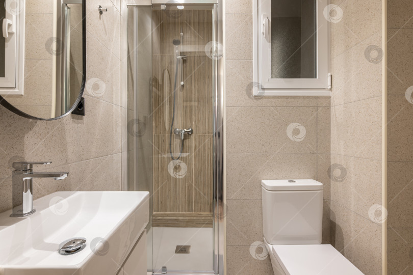 Скачать Красивая, свежая и удобная ванная комната для водных процедур, утренних и вечерних упражнений. Комната в бежевых тонах с большой раковиной, красивым зеркалом, стеклянной душевой кабиной и белым туалетом. фотосток Ozero