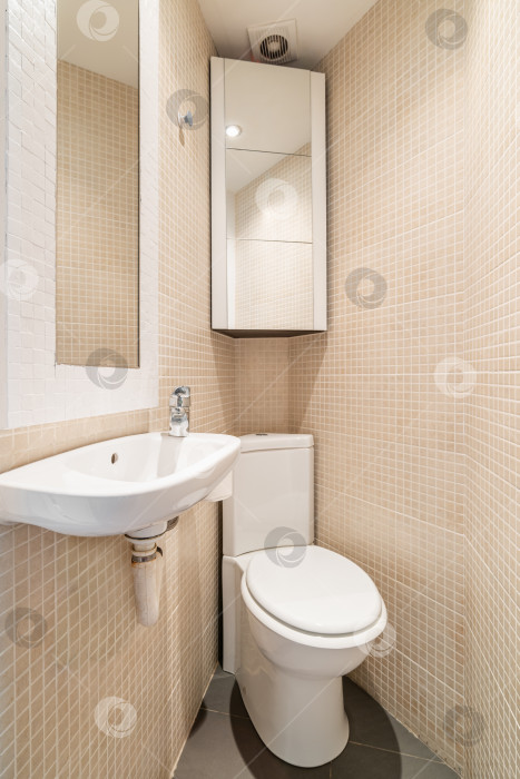 Скачать Вид на туалет в углу компактной конической ванной комнаты с бежевой мозаичной плиткой, зеркалом, унитазом, раковиной и настенным шкафом. Концепция дизайнерских решений в замкнутом пространстве фотосток Ozero