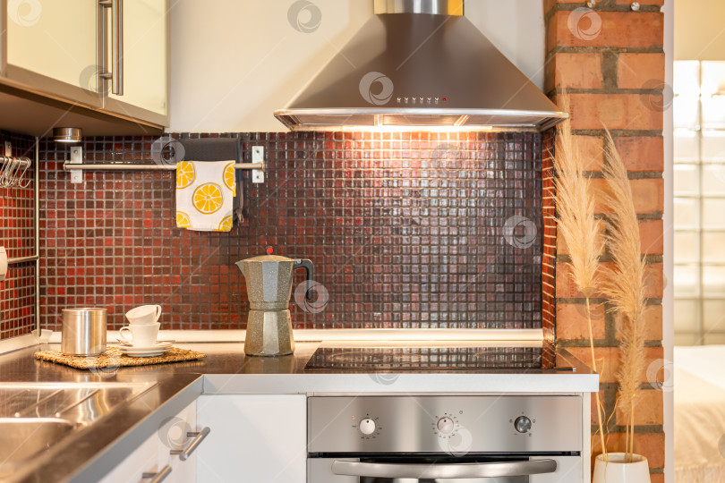 Скачать Вид на кухню с плитой, раковиной и вытяжкой, отделанную стильной бордовой мозаичной плиткой и белыми дверцами шкафчиков. Концепция стильного и комфортного кухонного интерьера фотосток Ozero