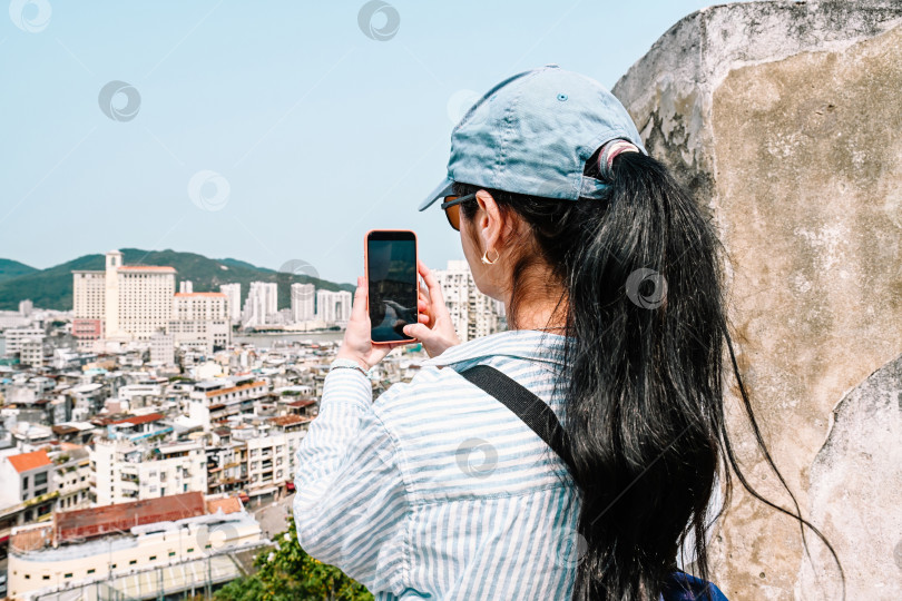Скачать Вид сзади на зрелую 50-летнюю русскую туристку в кепке, фотографирующую панораму города на телефон в крепости Монте, внесенной в список Всемирного наследия ЮНЕСКО, в Макао, Китай. фотосток Ozero