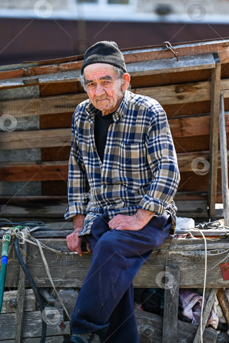 Скачать Бедный русский 77-летний пожилой мужчина сидит на улице и смотрит в камеру, демонстрируя концепцию бедности в сельской местности фотосток Ozero