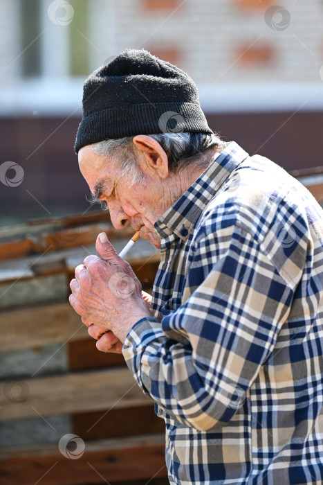 Скачать 77-летний пожилой мужчина из России прикуривает сигарету зажигалкой на улице в бедной деревне, демонстрируя вредную привычку и пристрастие к табаку фотосток Ozero