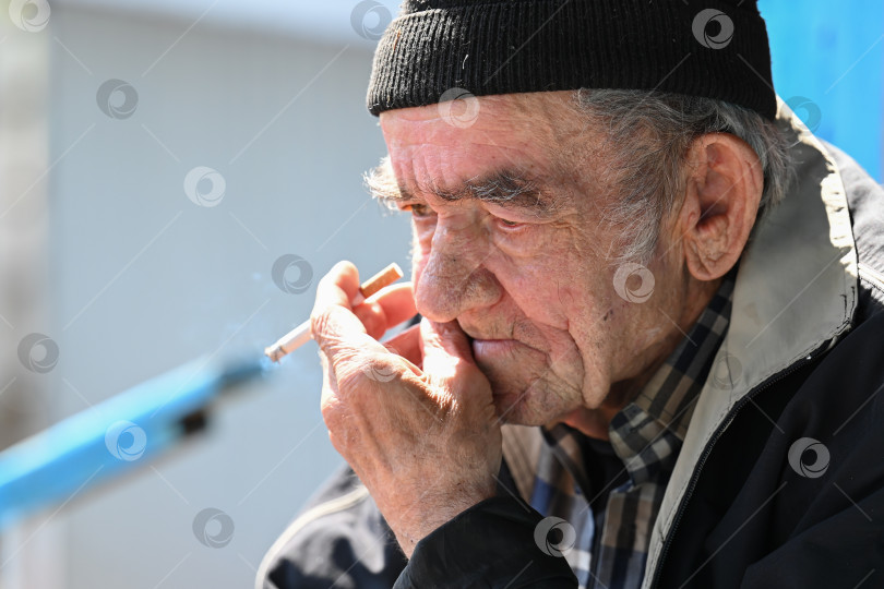Скачать Грустный русский 77-летний пожилой мужчина курит сигарету с задумчивым выражением лица в бедной деревне на открытом воздухе в России фотосток Ozero