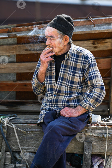 Скачать Бедный русский 77-летний пожилой мужчина сидит на улице и курит сигарету, демонстрируя вредную привычку и бедность в сельской местности фотосток Ozero