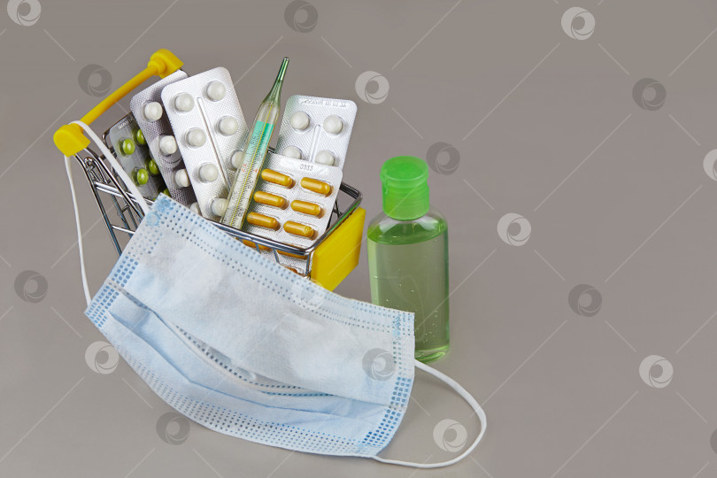 Скачать Таблетки, медикаменты, термометр, медицинская маска и дезинфицирующий гель в тележке супермаркета на сером фоне. фотосток Ozero