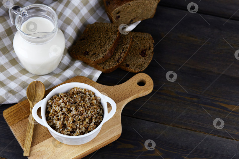 Скачать Гречневая каша в белой миске с ложкой, кувшин с молоком, нарезанный хлеб на темном деревянном фоне. фотосток Ozero