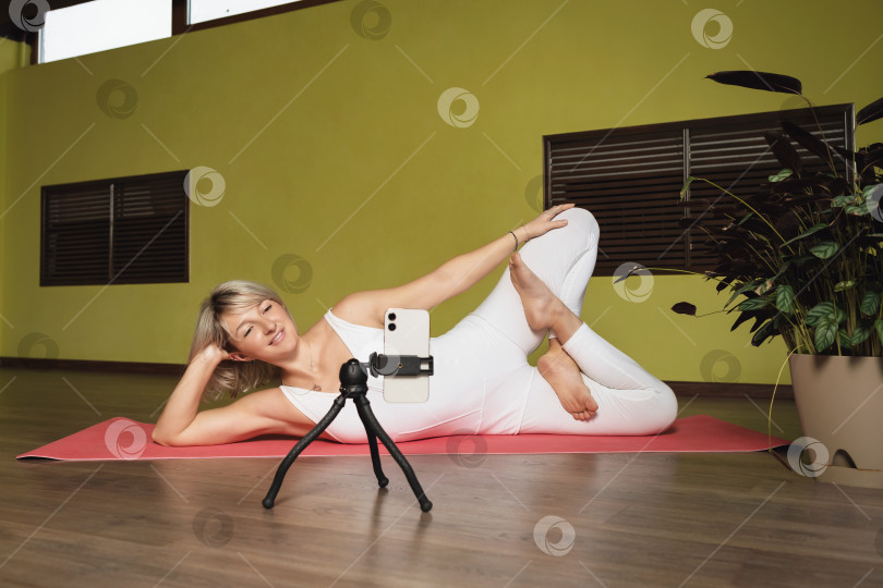 Скачать Женщина, ведущая здоровый образ жизни, ведет стримы для своей аудитории, лежит на коврике в спортивной одежде и выполняет позу лотоса, проводит онлайн-тренировку в студии фотосток Ozero