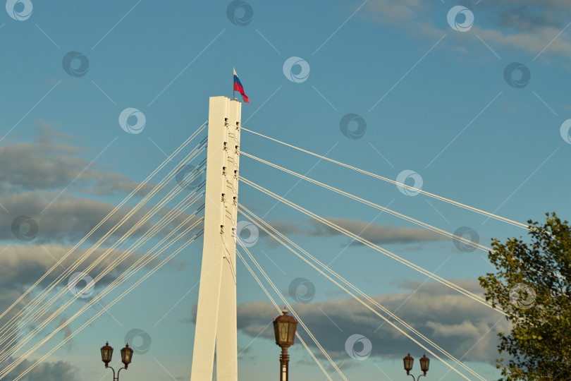 Скачать Тюмень, Россия - 6 сентября 2026 года: Верхняя часть белого пилона Моста влюбленных с развевающимся российским флагом на фоне голубого облачного неба и уличных фонарей фотосток Ozero