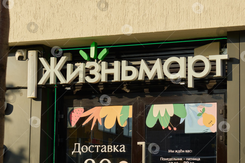 Скачать Тюмень, Россия - 6 сентября 2026 года: Неоновая вывеска продуктового магазина "Жизнмарт" крупным планом над стеклянной входной дверью на фасаде современного жилого дома фотосток Ozero