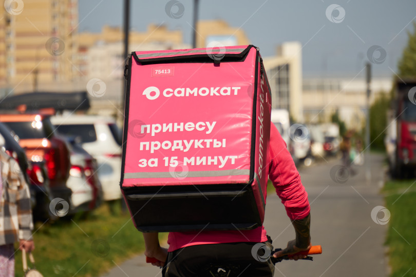 Скачать Тюмень, Россия - 6 сентября 2026 года: Вид сзади на курьера службы доставки еды "Самокат", едущего на велосипеде по городскому тротуару с большим розовым термо-рюкзаком в руках. фотосток Ozero
