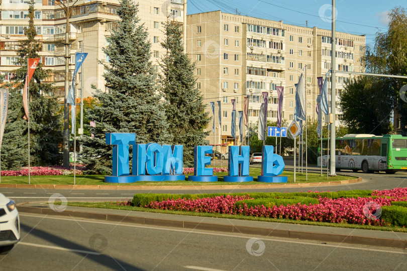Скачать Тюмень, Россия - 6 сентября 2026 года: Большие синие буквы с надписью "Тюмень" установлены на кольцевой развязке с цветочной клумбой, где проезжают городские автобусы и жилые здания фотосток Ozero