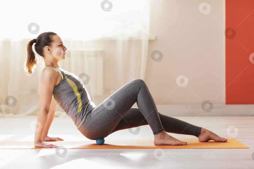 Скачать Женщина в спортивной одежде выполняет миофасциальный массаж ягодичных мышц мячом, сидя на коврике в студии фотосток Ozero