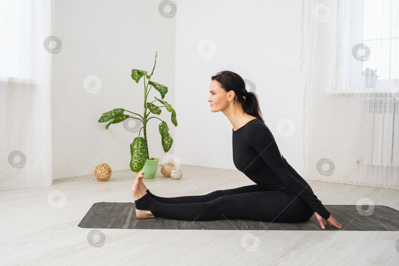 Скачать Женщина в черной спортивной одежде занимается йогой, выполняет наклон вперед, тренируется в комнате, сидя на коврике фотосток Ozero