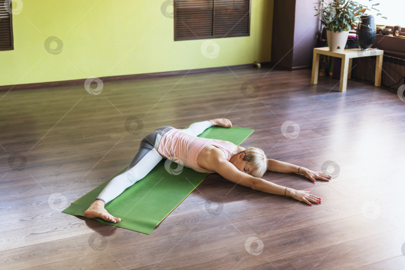 Скачать Женщина в спортивной одежде, практикующая йогу, выполняет упражнение самоканасана, поперечный шпагат с наклоном туловища вперед, лежит на коврике в студии фотосток Ozero