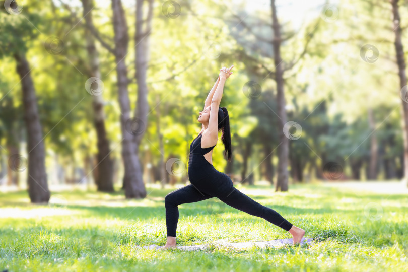 Скачать Молодая беременная женщина в черном спортивном комбинезоне практикует йогу, выполняет упражнение вирабхадрасана, позу воина, тренируется в парке теплым летним утром, стоя на коврике фотосток Ozero