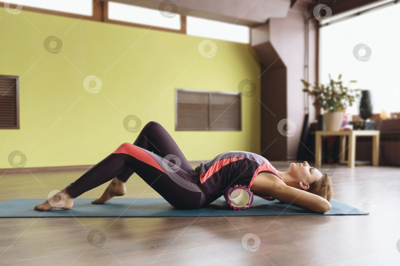 Скачать Женщина в спортивной одежде, ведущая здоровый образ жизни, выполняет миофасциальную релаксацию гиперактивных мышц спины с помощью массажного ролика, лежит на коврике в комнате фотосток Ozero