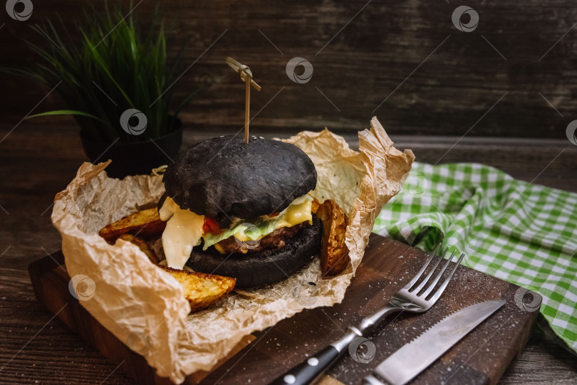 Скачать Черный бургер с сочной котлетой и картофелем по-деревенски в интерьере домашней кухни, завернутый в крафт-бумагу. Черная булочка для бургера скрепляется бамбуковой шпажкой. Домашняя кухня. фотосток Ozero