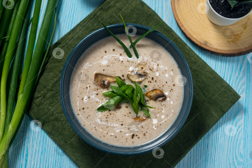 Скачать Грибной суп-пюре с кусочками шампиньонов и мелко нарезанным зеленым луком в темно-синей миске на зеленой льняной салфетке. Вид сверху. Деревянные ложки, синяя деревянная столешница. фотосток Ozero