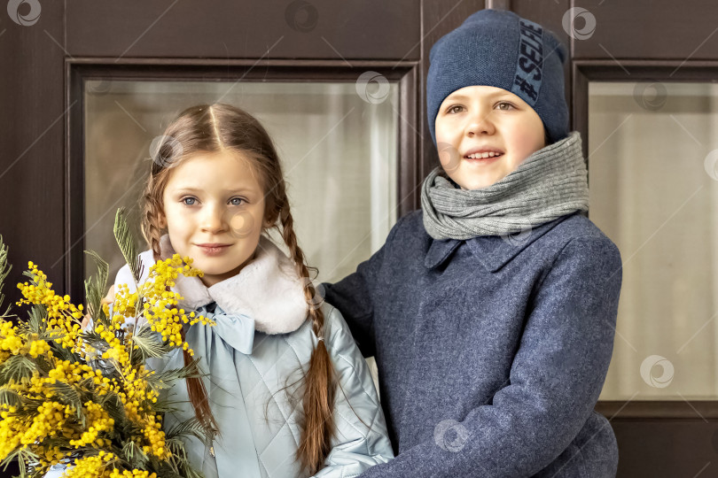 Скачать Портрет пары мальчика и девочки школьников в весенних пальто с букетом мимозы в руках в парке. Весна, Международный женский день 8 марта. фотосток Ozero