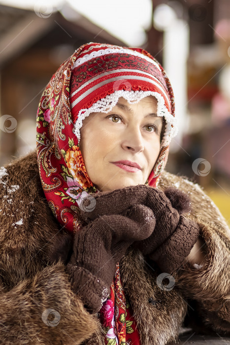 Скачать Славянская женщина в национальном цветном шарфе, шубе и теплых варежках на крыльце деревянного дома. Зима, снегопад фотосток Ozero