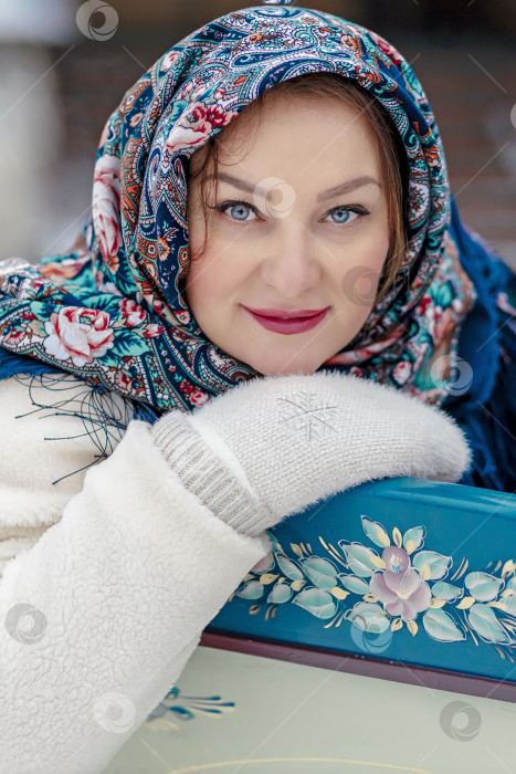 Скачать Славянская женщина в национальном цветном шарфе, шубе и теплых варежках сидит на деревянных санях. Зима, снегопад фотосток Ozero