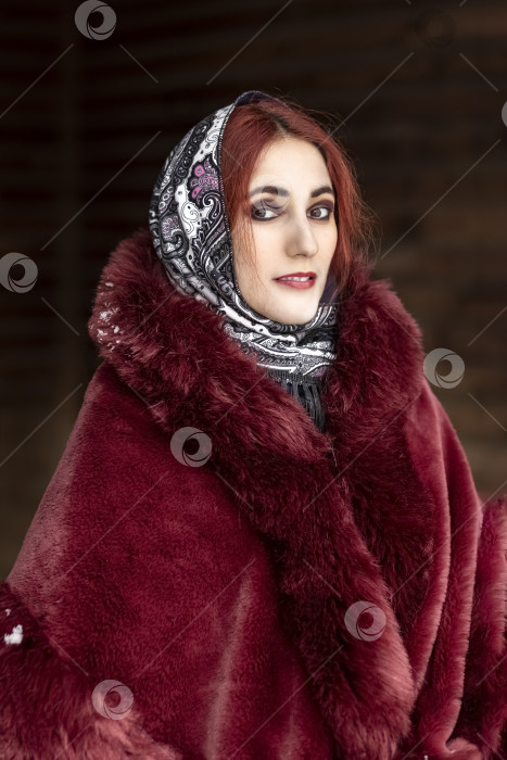 Скачать Славянская женщина в национальном цветном шарфе, шубе и теплых варежках на крыльце деревянного дома. Зима, снегопад фотосток Ozero