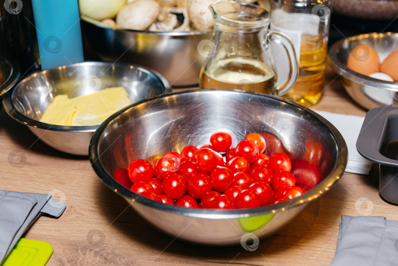 Скачать Приготовление блюда, сырые ингредиенты на столе, очищенные помидоры черри в металлической миске на деревянном столе, кувшин растительного масла и кусочек сливочного масла фотосток Ozero