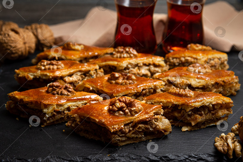 Скачать Медовая пахлава с грецкими орехами на черном грифельном столе. Порции восточных сладостей. Кусочки турецкого сахара. Чай, налитый в стакан армуду на заднем плане. фотосток Ozero