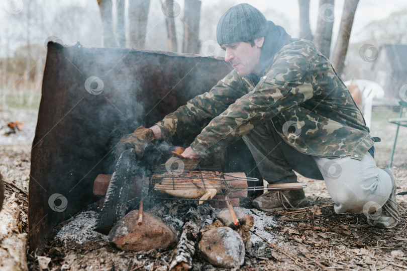 Скачать молодой человек-выживальщик готовит блюда из куриного мяса, обжаренные на гриле на тлеющих углях или тлеющих угольках от костра на земле. барбекю в походных условиях. сельская местность и дикая деревенская жизнь фотосток Ozero