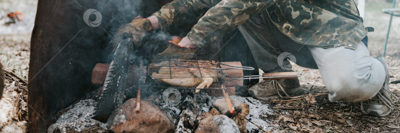 Скачать молодой человек-выживальщик готовит блюда из куриного мяса, обжаренные на гриле на тлеющих углях или тлеющих угольках от костра на земле. барбекю в походных условиях. сельская местность и дикая деревенская жизнь фотосток Ozero