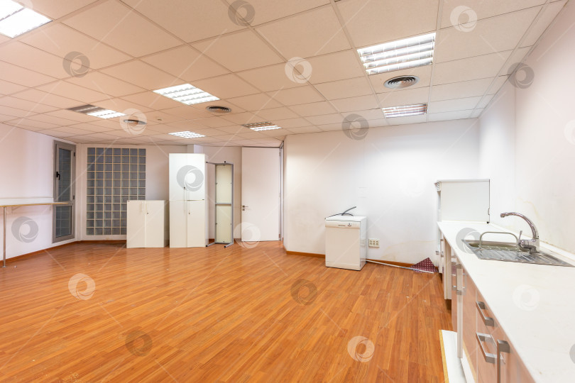 Скачать Старая пустая комната с белой кухонной мебелью и раковиной, которая раньше служила офисной столовой для сотрудников. Просторное и светлое помещение, оборудованное как корпоративная столовая, нуждается в новом ремонте. фотосток Ozero