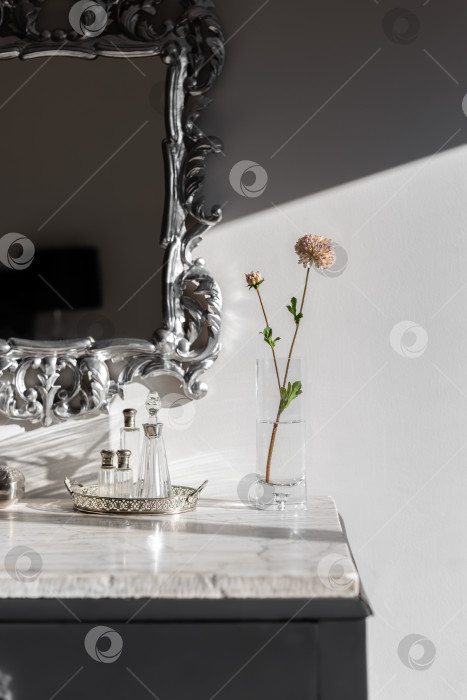 Скачать Часть туалетного столика с вазой и цветком, ярко освещенными солнечным светом на столешнице из белого мрамора. Зеркало в серебряной раме с красивыми фигурными вензелями. На столе стоят пустые флаконы из-под ароматных духов. фотосток Ozero
