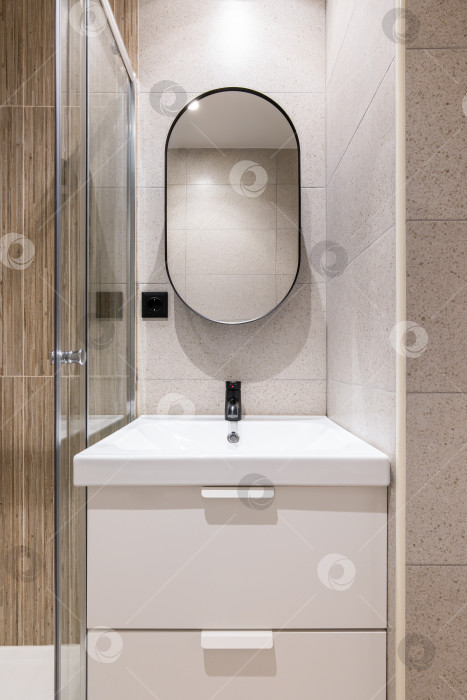 Скачать Крупным планом ниша в ванной комнате с ярким искусственным освещением, с белой раковиной из цельной керамики на туалетном столике с двумя выдвижными ящиками. Большое современное овальное зеркало на стене над раковиной. фотосток Ozero