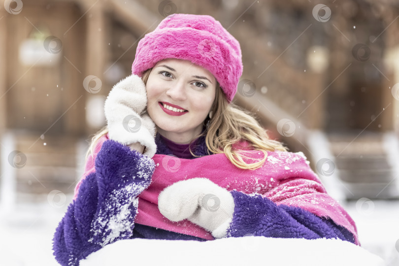 Скачать Портрет улыбающейся молодой женщины в ярко-розовом шарфе и шляпе, белых варежках и фиолетовой шубе на открытом воздухе в городском парке зимой. фотосток Ozero