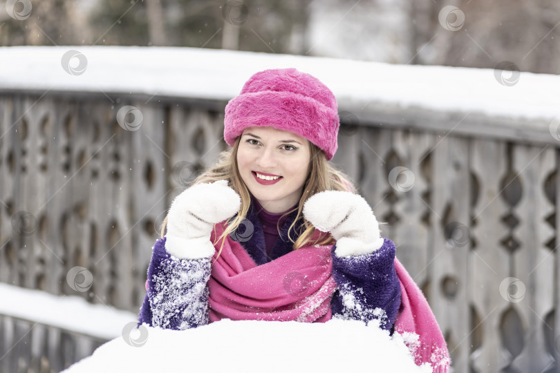 Скачать Портрет улыбающейся молодой женщины в ярко-розовом шарфе и шляпе, белых варежках и фиолетовой шубе на открытом воздухе в городском парке зимой. фотосток Ozero