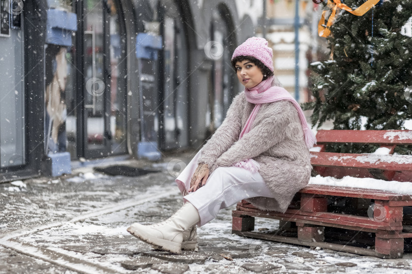 Скачать Портрет улыбающейся стильной молодой женщины в теплой одежде пастельных тонов на улице в городе зимой.Снегопад, холодная погода фотосток Ozero
