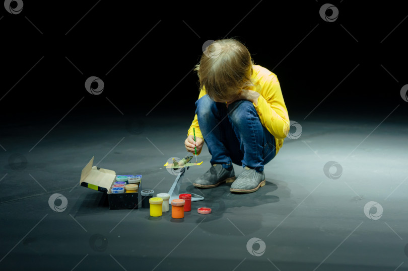 Скачать откровенный маленький шестилетний мальчик рисует или раскрашивает модель самолета гуашевыми красками, сидя на корточках на полу студии, освещенной светом фотосток Ozero