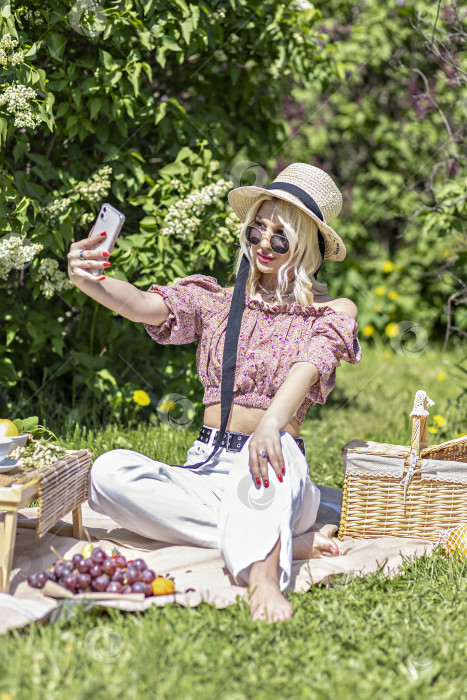Скачать Портрет молодой светловолосой женщины в шляпе и солнцезащитных очках на пикнике в парке.Делает селфи на смартфон во время летних каникул.Видеочат фотосток Ozero