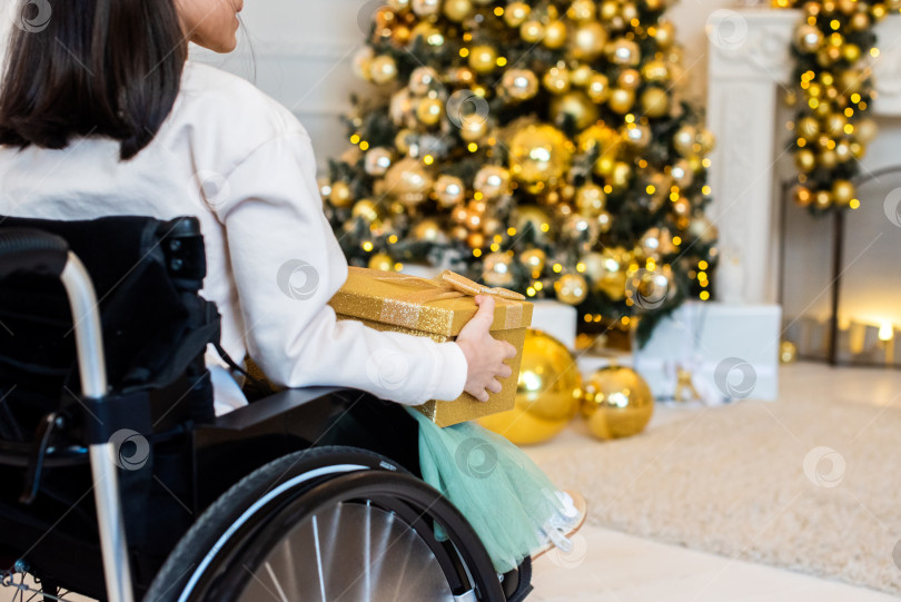 Скачать маленькая девочка в инвалидном кресле возле рождественской елки. инвалид, держащий желтый, золотой новогодний подарок фотосток Ozero