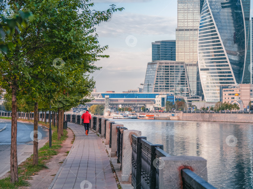 Скачать Утренняя фитнес-пробежка по набережной в Москве. Мужчина в красной спортивной ветровке бежит по пустынной улице ранним утром фотосток Ozero