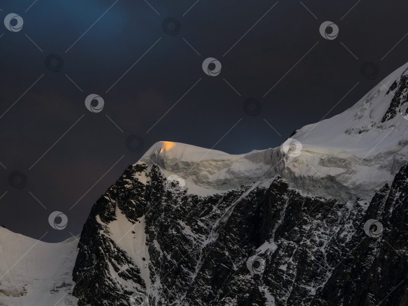 Скачать Темный горный пейзаж с огромной снежной горой, освещенной рассветным солнцем среди темных облаков. Потрясающий альпийский пейзаж с высокой горной вершиной на закате или восходе солнца. Большой ледник на вершине в оранжевом свете. фотосток Ozero