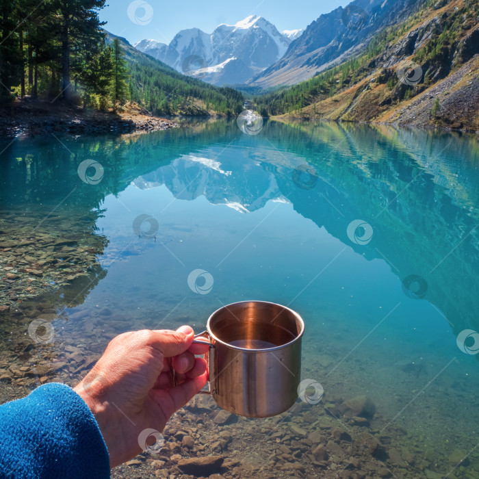 Скачать Рука, держащая походную кружку с чистой водой. Чистая питьевая вода в голубом горном озере. Чистая вода в туристическом стаканчике на фоне озера и заснеженных гор. фотосток Ozero