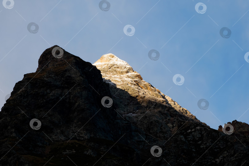 Скачать Контрастирующие горы на закате. Вечерний горный пейзаж со снежной скалой в золотистом солнечном свете. Природный фон прогулки по скалистым горам с острыми скалами и голубым небом. фотосток Ozero