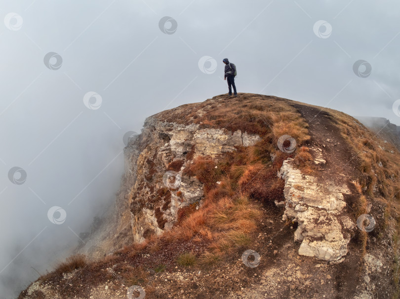 Скачать Силуэт человека над туманными облаками, утренний холмистый пейзаж. Одинокая скала с человеком-путешественником над облаками туманным утром на Кавказе. фотосток Ozero