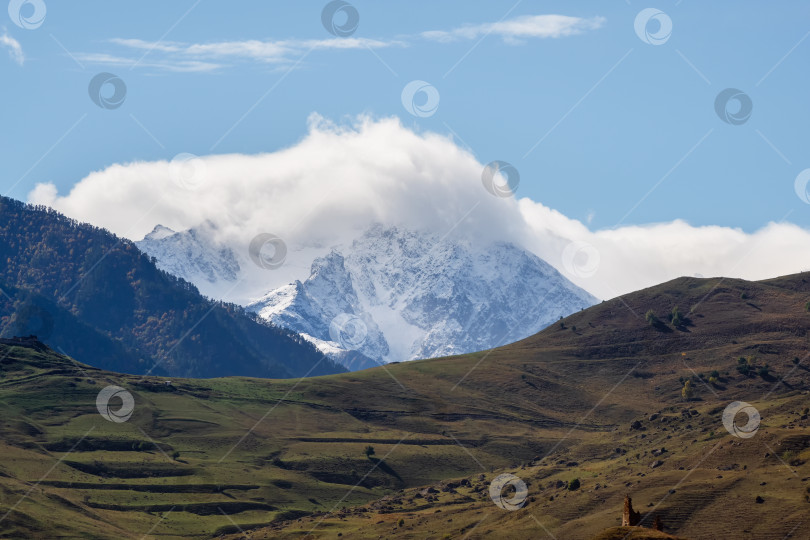 Скачать Живописный пейзаж с осенней горной долиной на фоне больших гор в низких облаках при утреннем солнечном свете. Яркий склон холма с видом на залитый солнечным светом высокий горный хребет в низких облаках. фотосток Ozero