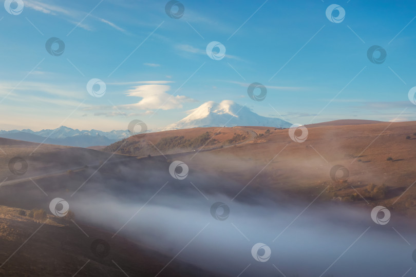 Скачать Мягкий фокус. Гора Эльбрус в густом утреннем тумане и на солнечном склоне. Мистический пейзаж с прекрасными заснеженными скалами в низких облаках. Прекрасный горный туманный пейзаж. фотосток Ozero